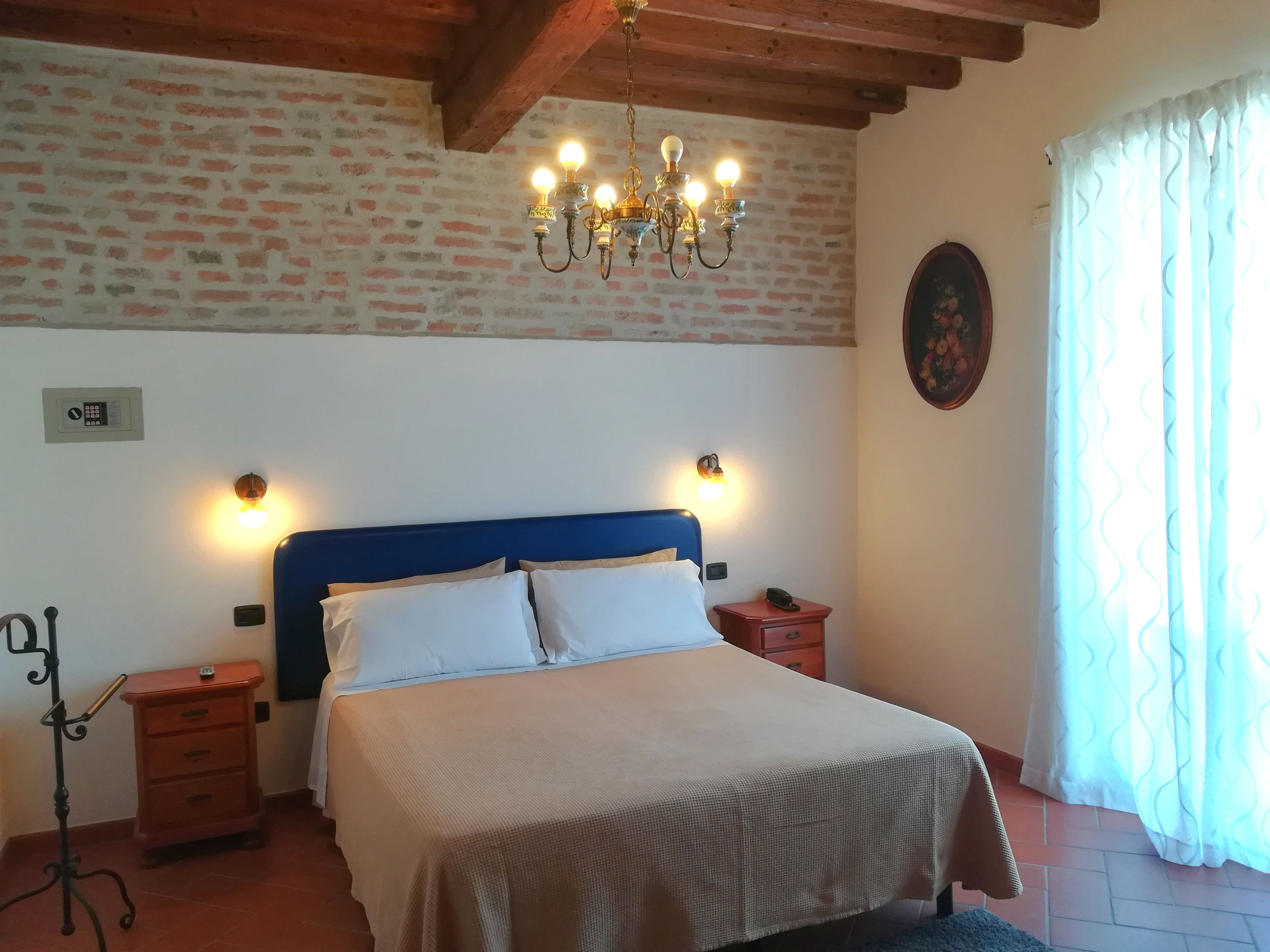 Une chambre avec un mur en briques, une tête de lit bleue, un lit avec une literie blanche, un lustre, des lampes de chevet, une table de chevet, une armoire en bois et une fenêtre avec des rideaux.