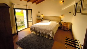 Un dormitorio con cama, vigas de madera en el techo, una mesita de noche con lámpara, un pequeño aparador de madera y vistas al exterior a través de una puerta de cristal.