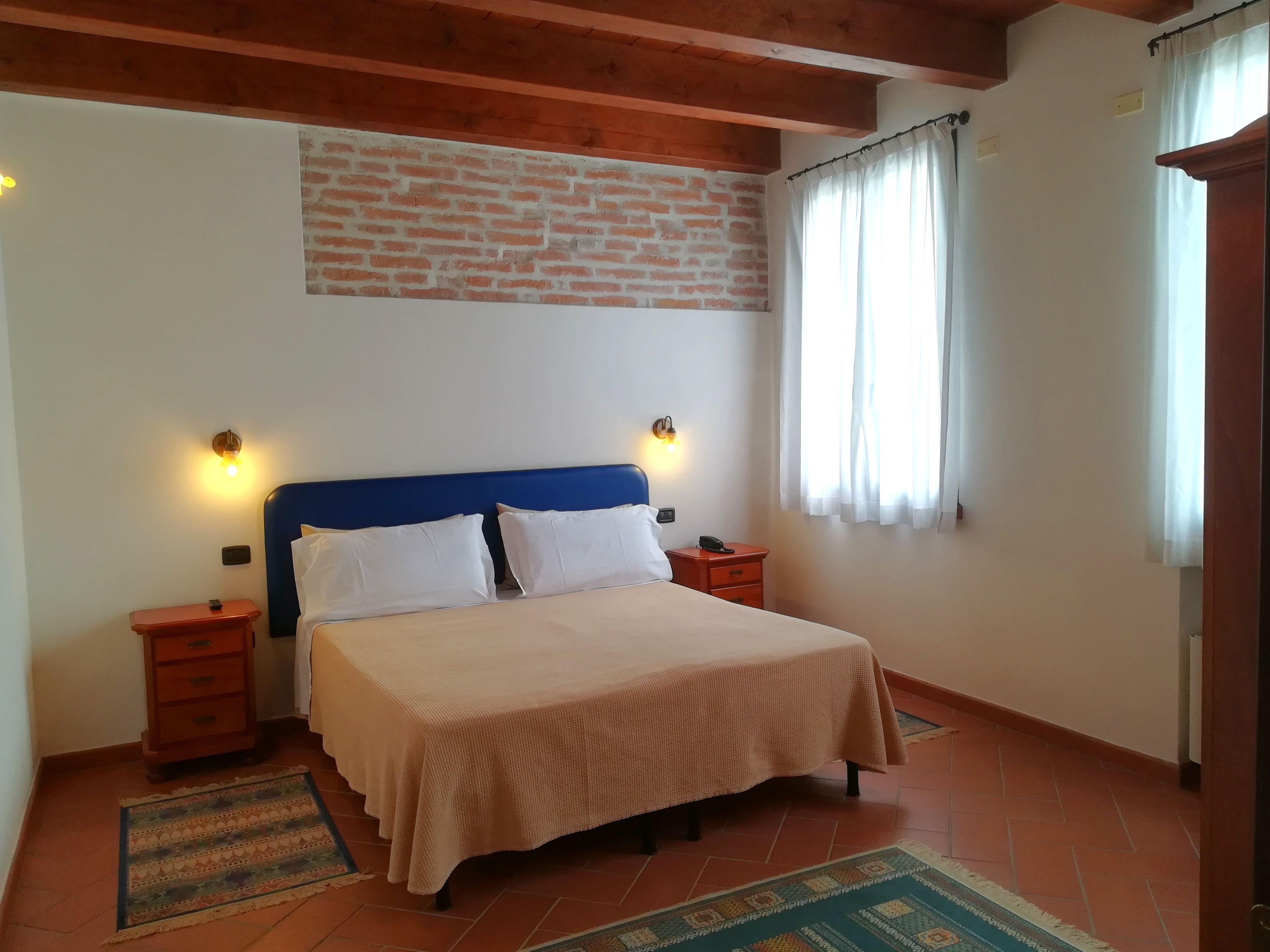 Un dormitorio con cama, dos mesitas de noche, una mesilla, pared de ladrillo, ventana con cortinas y techo de madera.