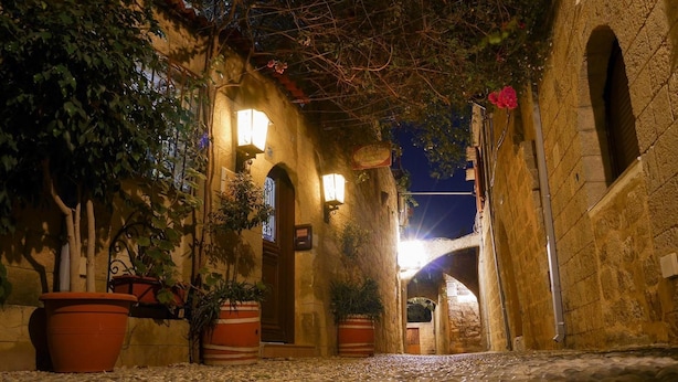 A cobblestone alleyway with stone walls, potted plants, and illuminated signs. - Sofia Pension (Rhodes)