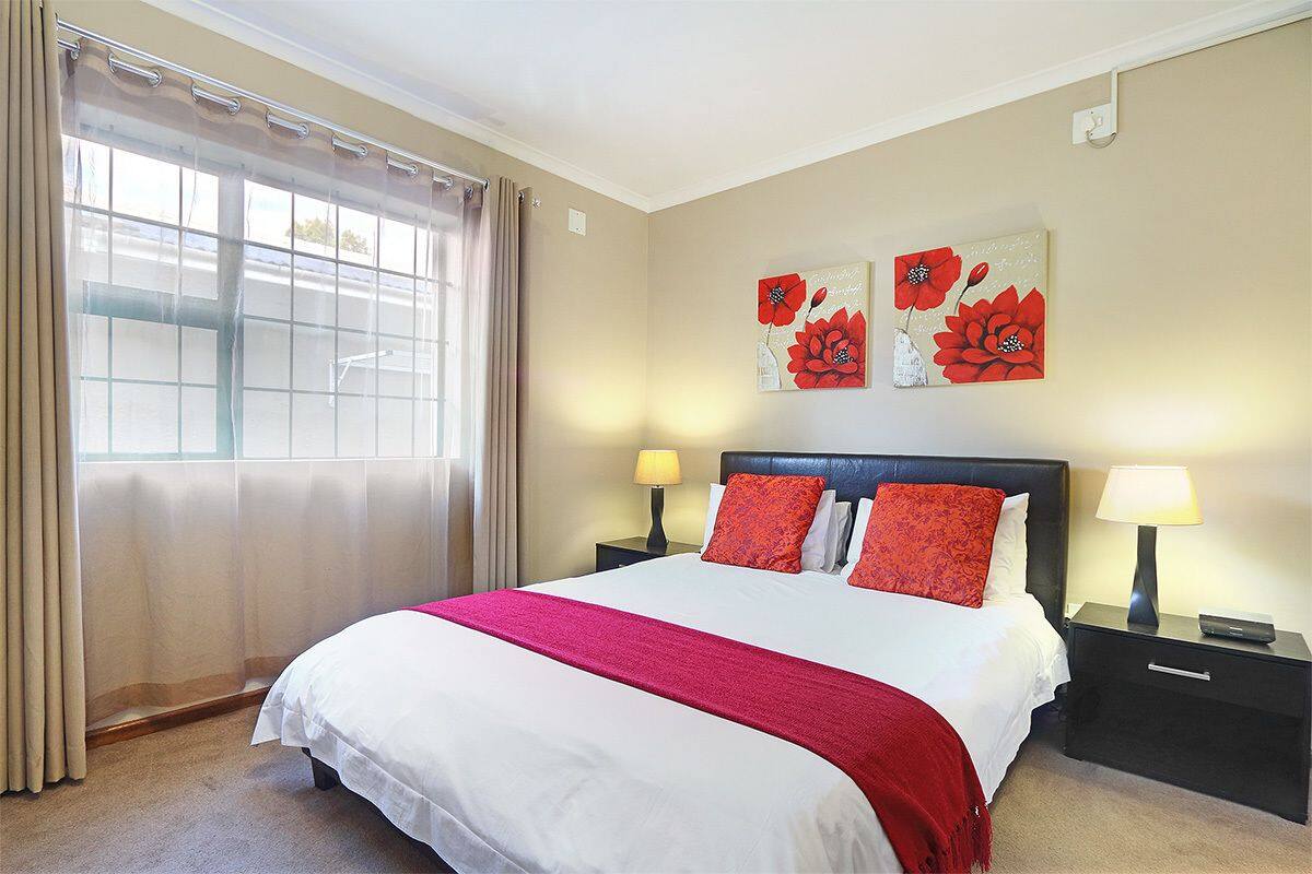 A hotel room with a large bed, two bedside tables with lamps, a window with curtains, and two wall-mounted paintings of red flowers.