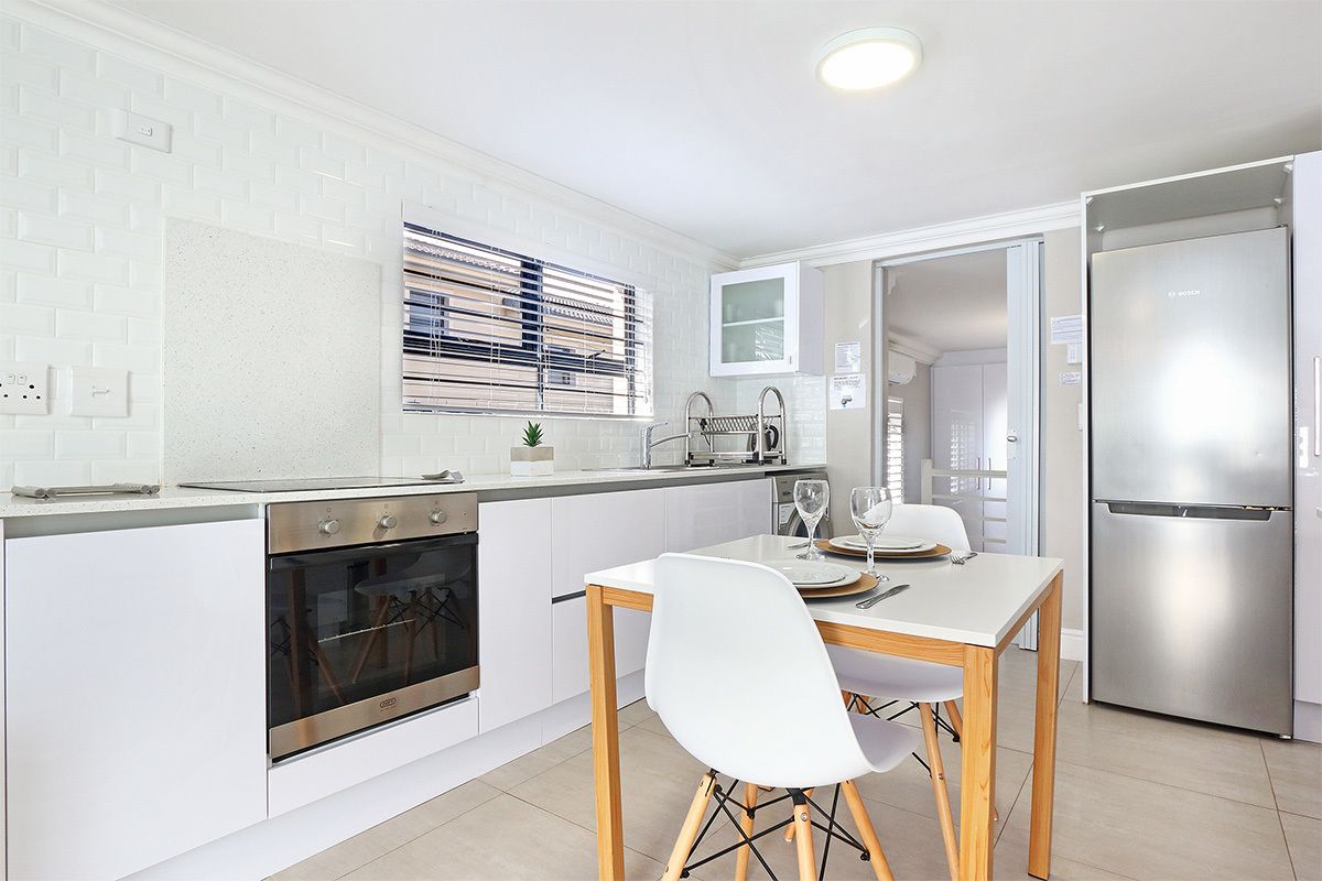 Una cocina moderna con armarios blancos, horno integrado, refrigerador de acero inoxidable y una zona de comedor con una silla y una mesa blancos.