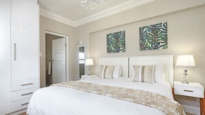 A bedroom with a large bed, white headboard, two nightstands with lamps, a wardrobe, and two abstract leaf-patterned wall art pieces. - Star Holiday Apartments (Cape Town)