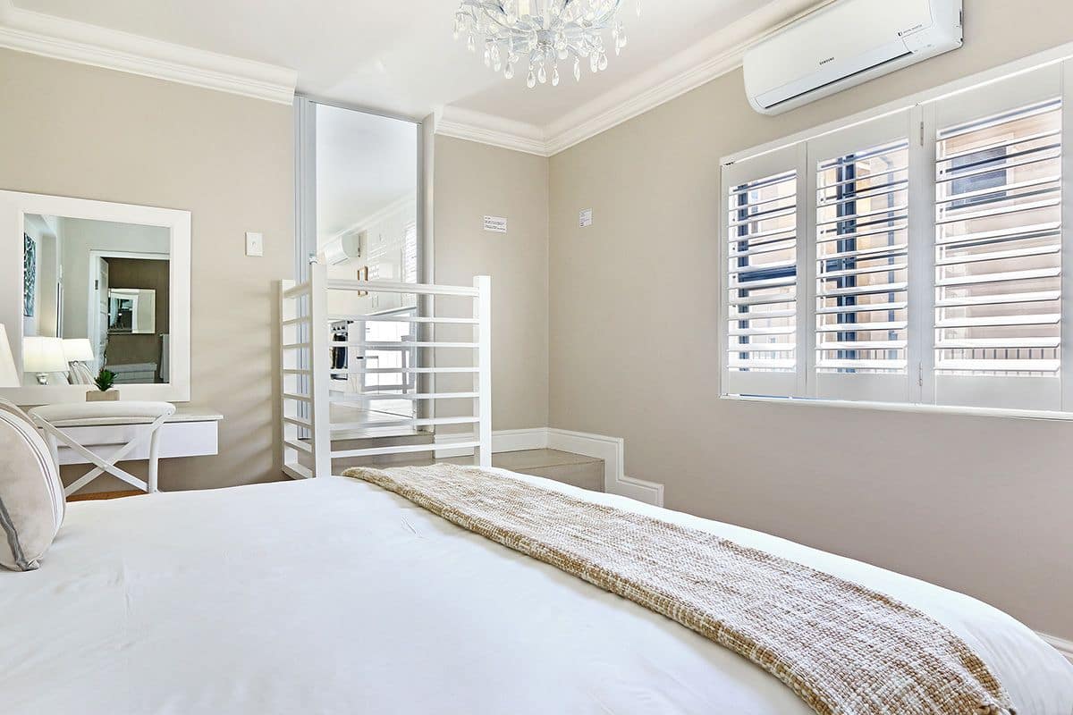 A bedroom with a bed, a desk, a mirror, and a window with blinds.