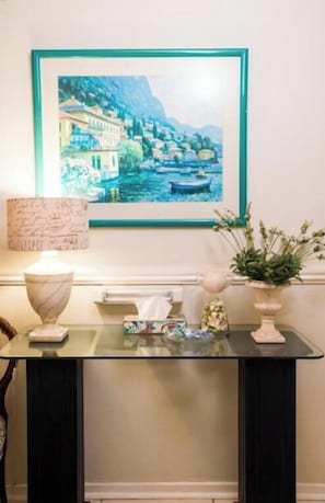 Uma mesa com tampo de vidro, uma luminária de mármore, um quadro de uma vila costeira, um vaso com flores e uma pequena planta em vaso.