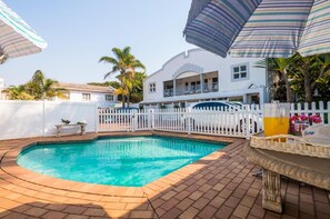 Piscina com cerca branca, guarda-sóis e uma mesa com bebidas.
