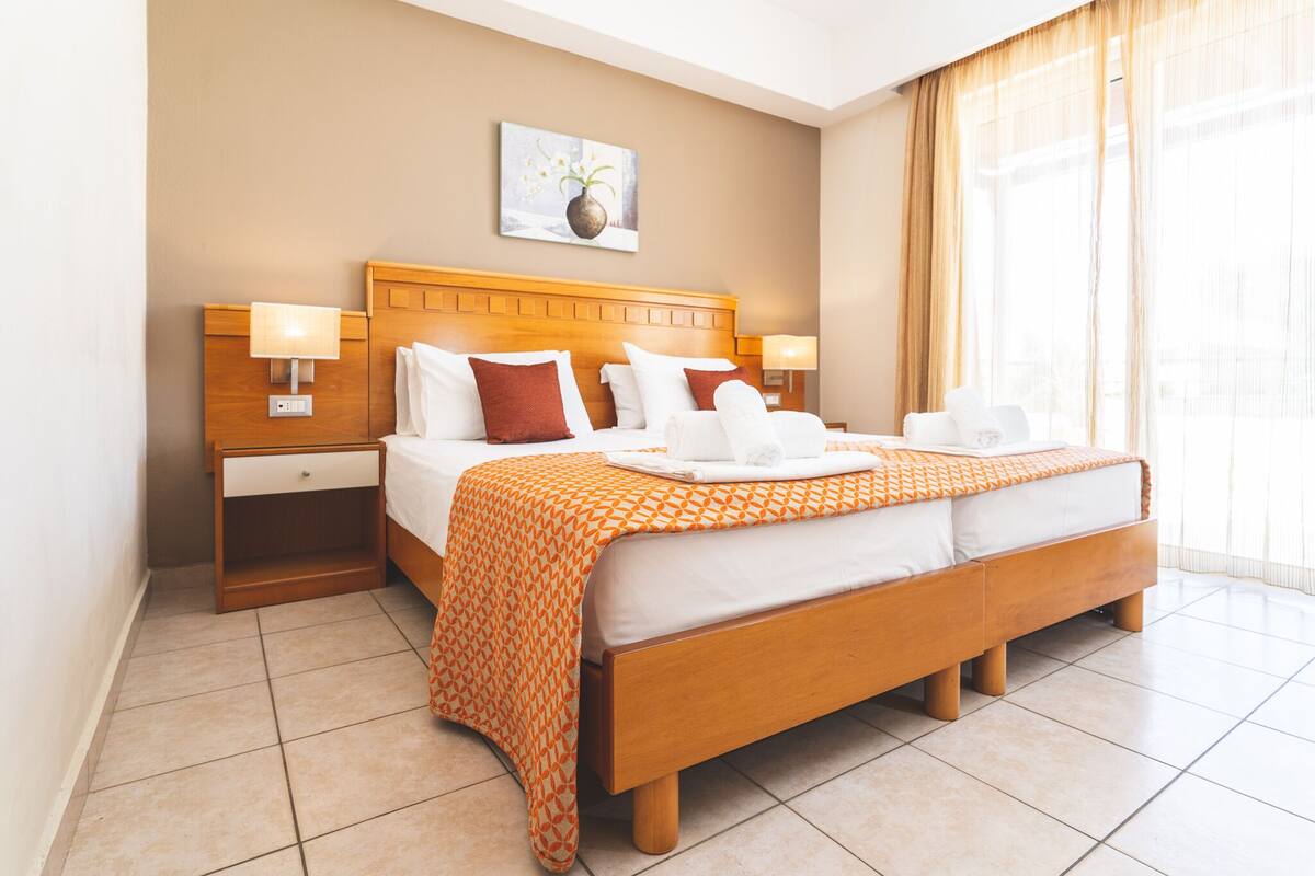 Une chambre d’hôtel avec un grand lit, deux tables de chevet, une petite table, une lampe fixée au mur et une fenêtre avec des rideaux.