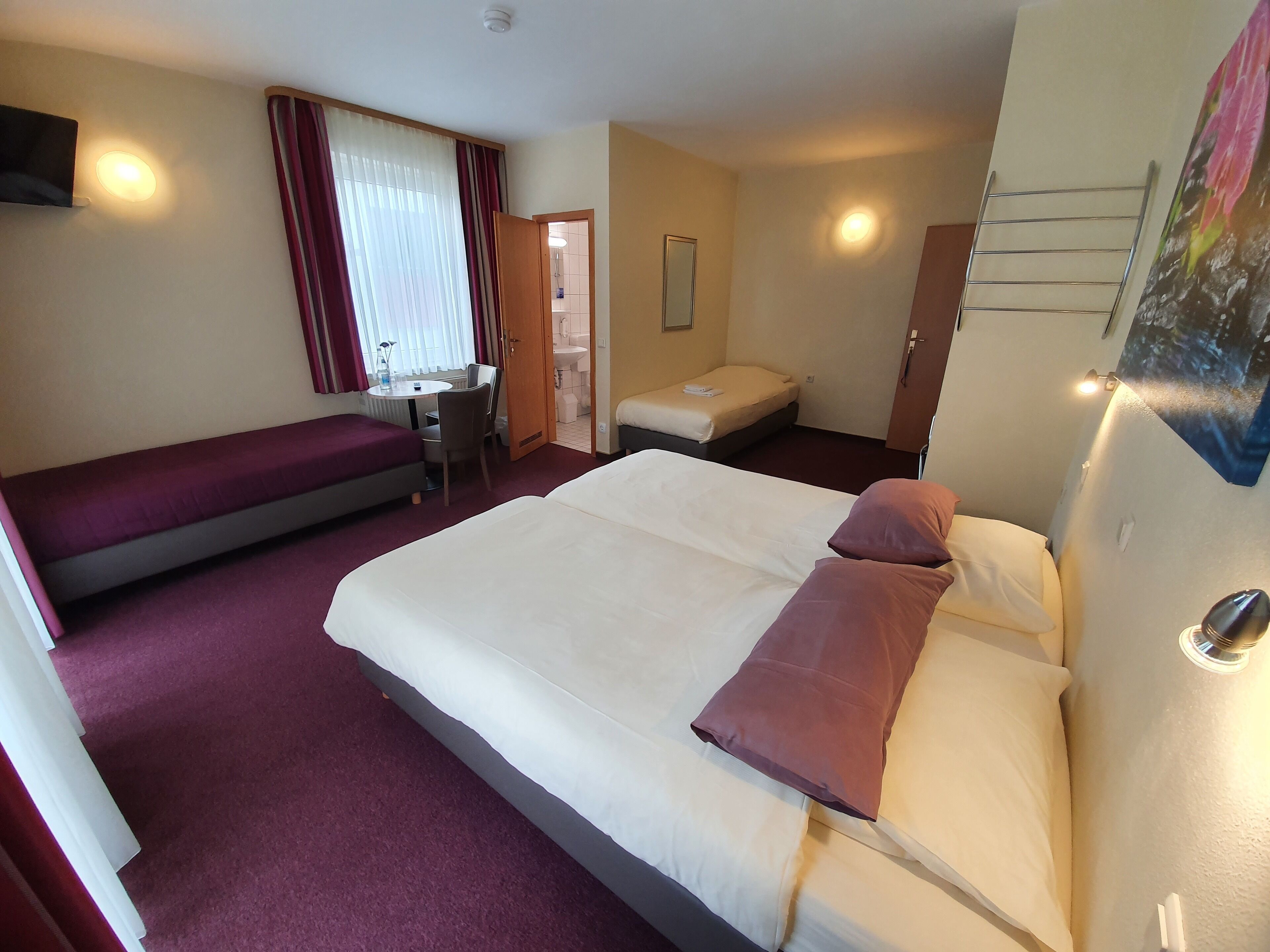 A hotel room with a large bed, a smaller bed, a desk with a chair, a window with curtains, and a bathroom visible through an open door.