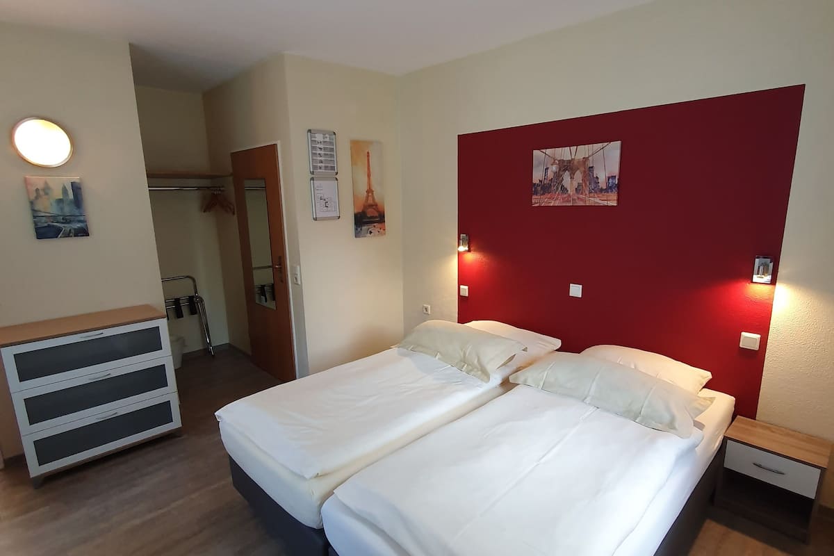 Un lit double avec une literie blanche, un mur d’accent rouge et une petite table de chevet avec une lampe.