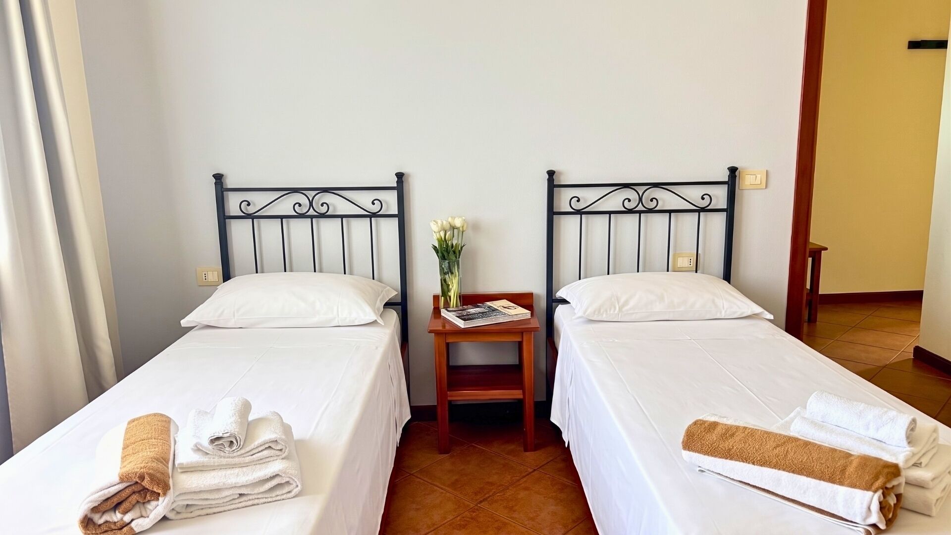 A hotel room with two single beds, a small table with a vase of flowers, and towels on the beds.