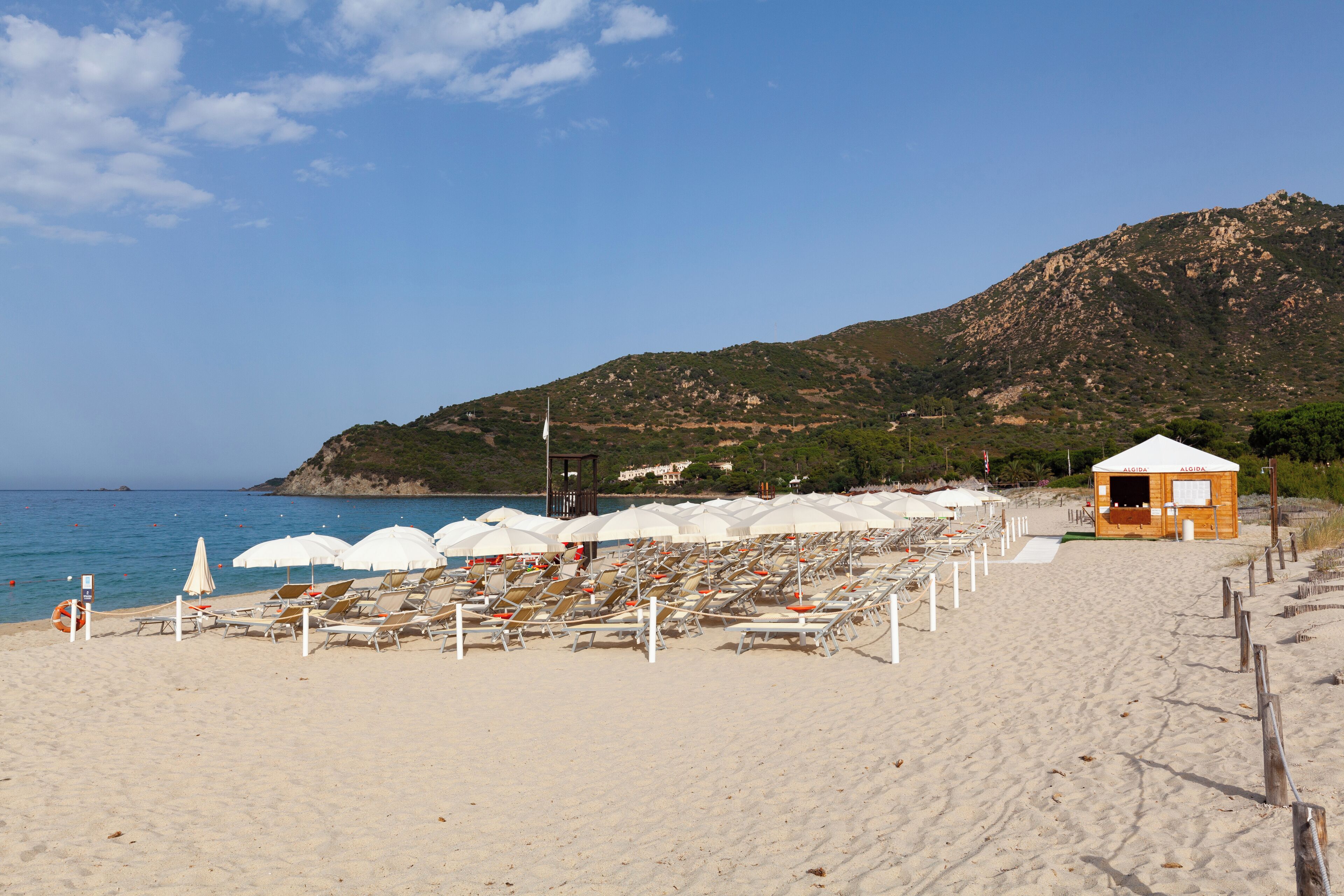 A beach with rows of sun loungers and umbrellas, a lifeguard tower, and a small wooden building labeled 'AQUA'.