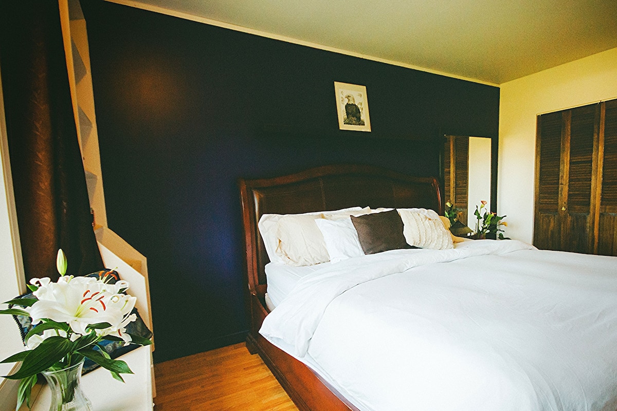 Ein Schlafzimmer mit einem hölzernen Bett, weißer Bettwäsche, einer Vase mit Blumen und einem gerahmten Bild an der Wand.