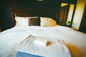 Una cama bien hecha con una colcha blanca, un plaid azul y una almohada blanca con detalles marrones. Una caja blanca con la etiqueta 'OKAY' está colocada sobre la cama.