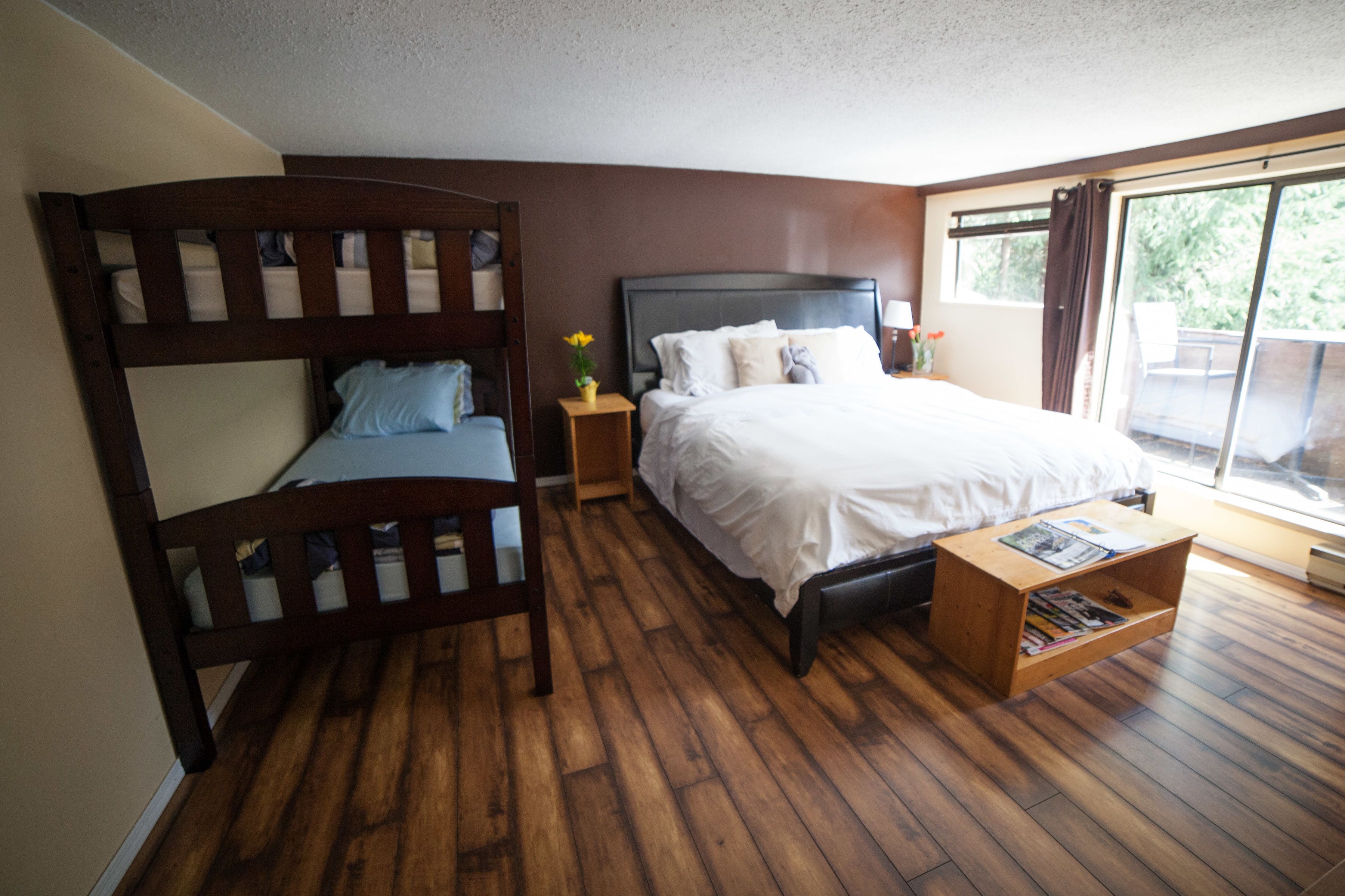 A bedroom with a bunk bed, a single bed, a wooden nightstand with a lamp, a bedside table with books, and a window with curtains.