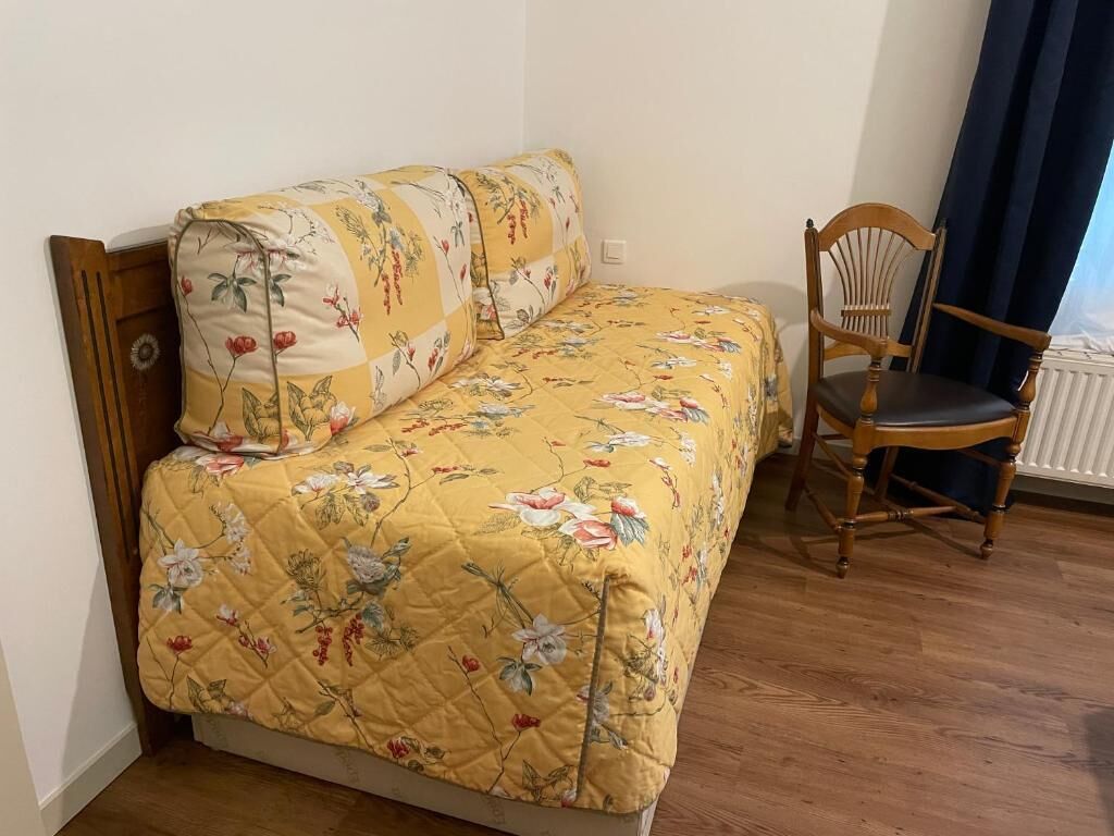 Un lit simple avec une couette à motifs floraux, une chaise en bois et un radiateur dans une pièce dont le sol est recouvert de planches de bois.