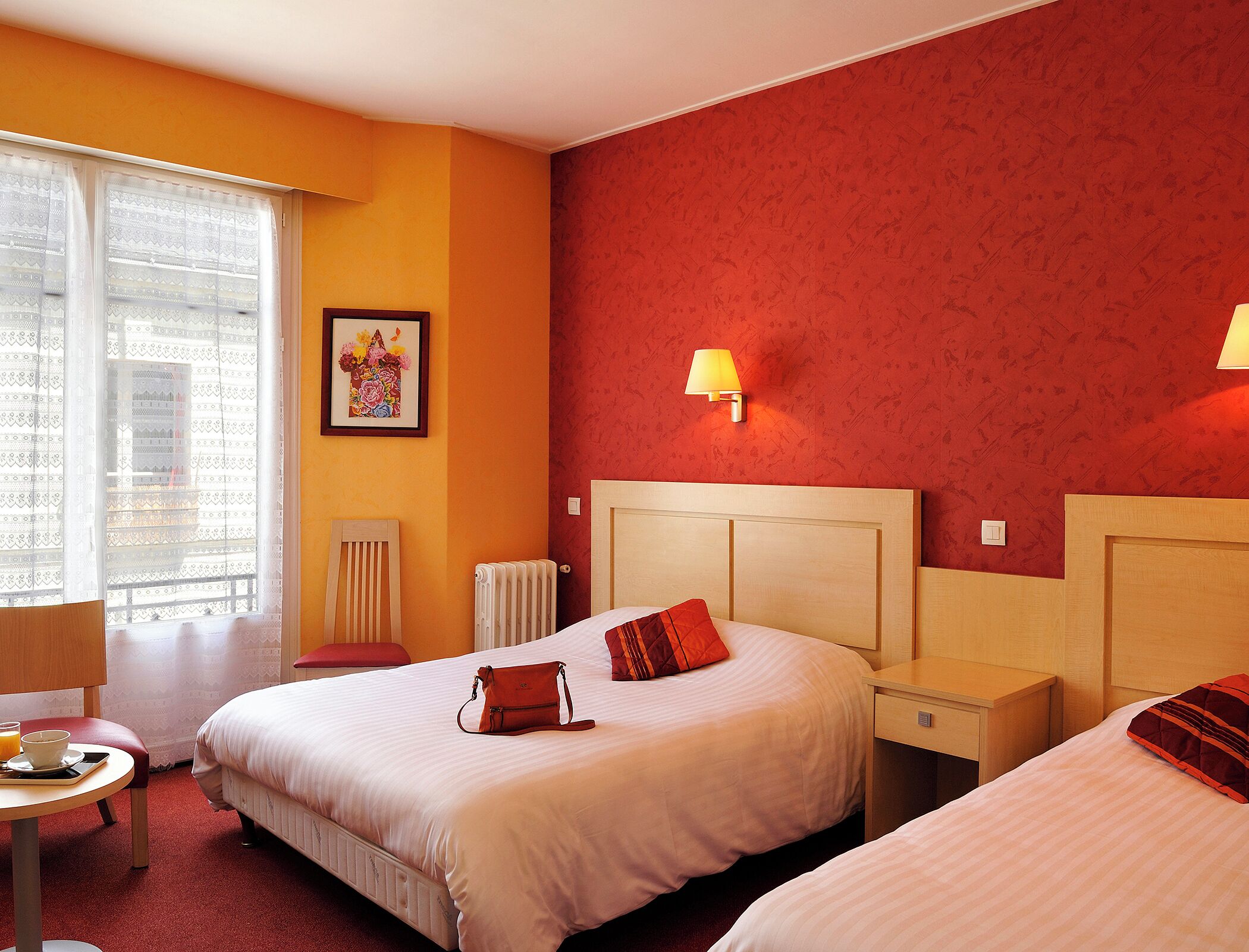 Habitación de hotel con dos camas, una ventana con cortinas translúcidas, un radiador, una mesita de noche con una lámpara y un cuadro enmarcado en la pared.