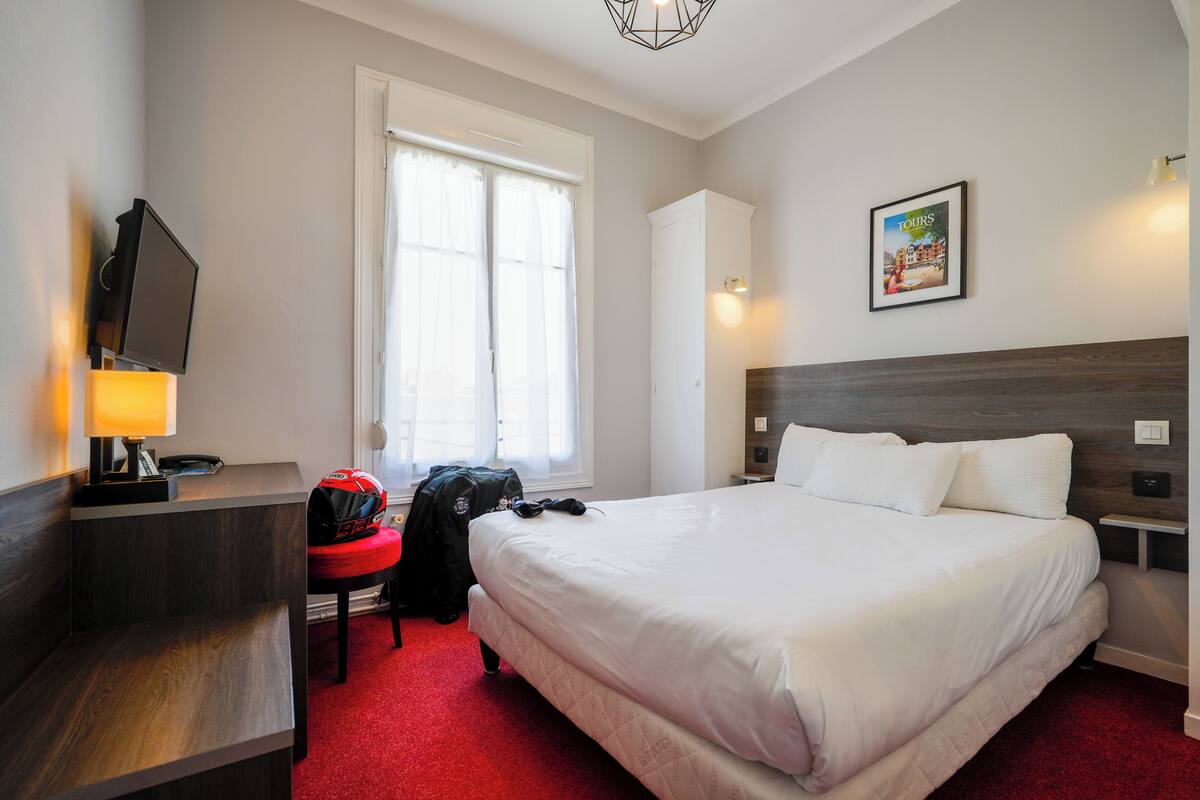 Une chambre d’hôtel avec un lit, un bureau doté d’une lampe, une chaise, une fenêtre et un tableau accroché au mur.