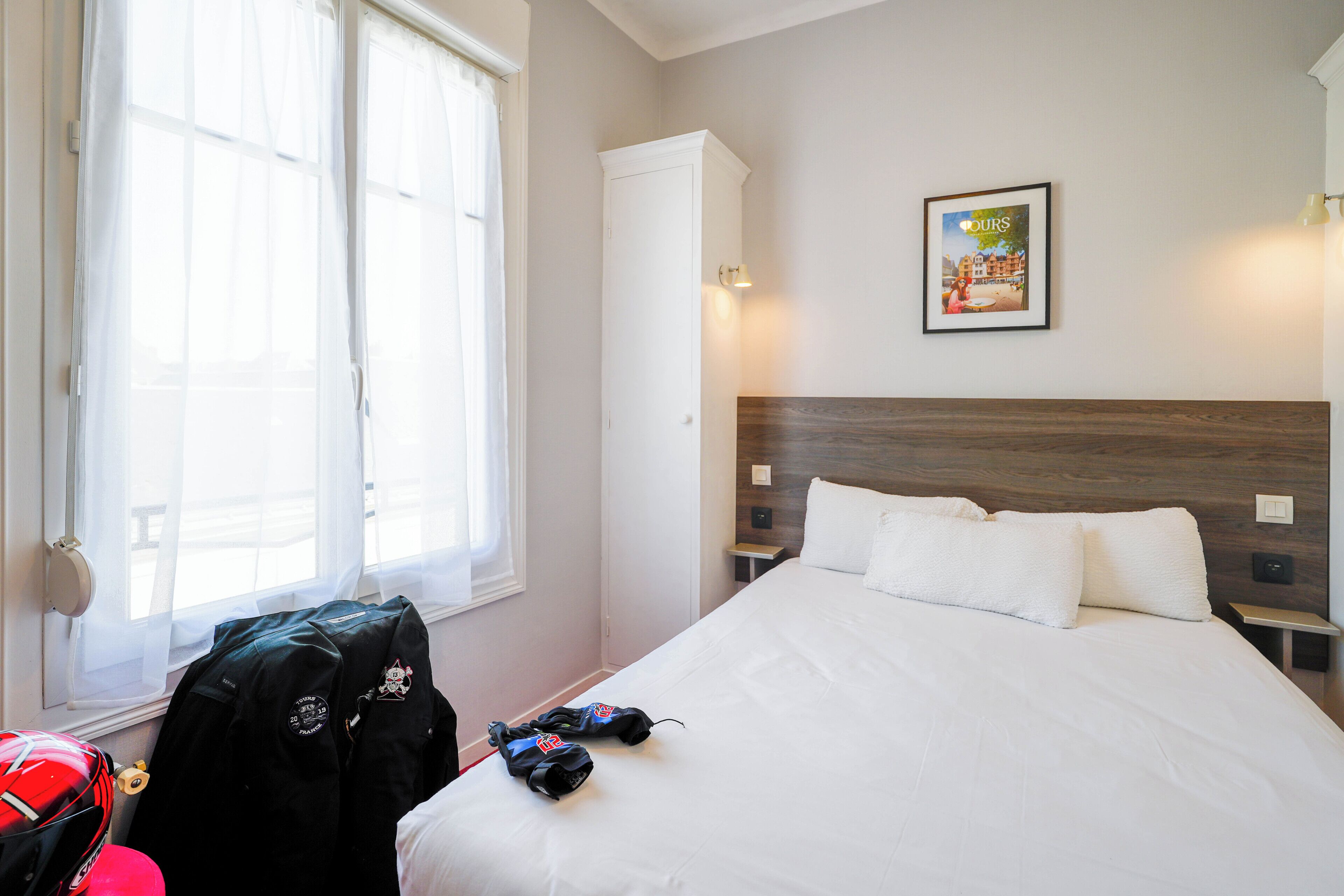 A hotel room with a bed, a window with sheer curtains, a framed picture on the wall, and a suitcase on the floor.