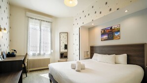 Quarto de hotel com uma cama grande, uma escrivaninha, uma cadeira, um radiador, uma janela com cortinas, um espelho e um quadro de uma paisagem urbana na parede.