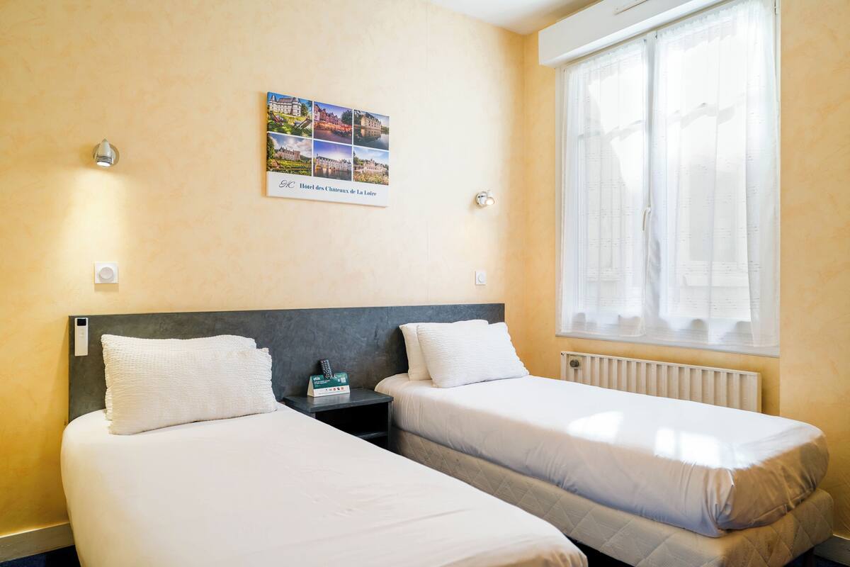 Une chambre d’hôtel avec deux lits, une table de chevet, une lampe fixée au mur, une fenêtre avec des rideaux et une affiche décorative sur le mur.