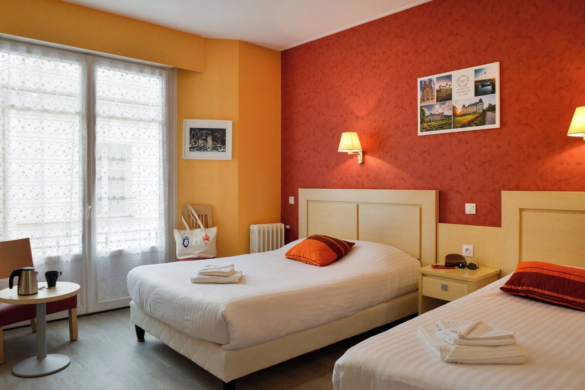Une chambre d’hôtel avec deux lits, une table avec une bouilloire, une chaise et une fenêtre avec des rideaux.