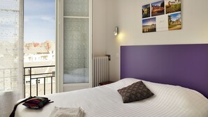 Quarto de hotel com uma cama grande, uma janela com vista para edifícios e uma parede decorada com quadros.