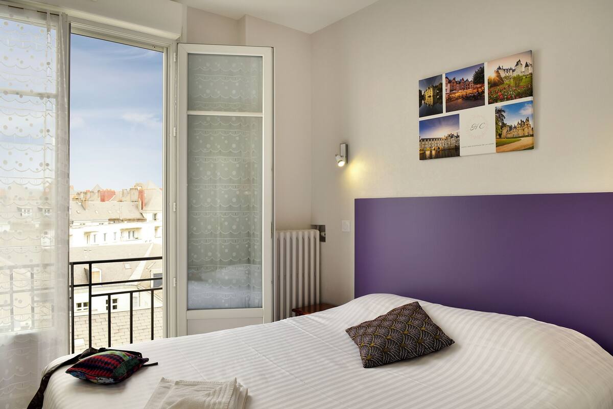 Une chambre d’hôtel avec un grand lit, une fenêtre donnant sur des immeubles et un mur décoré de tableaux.