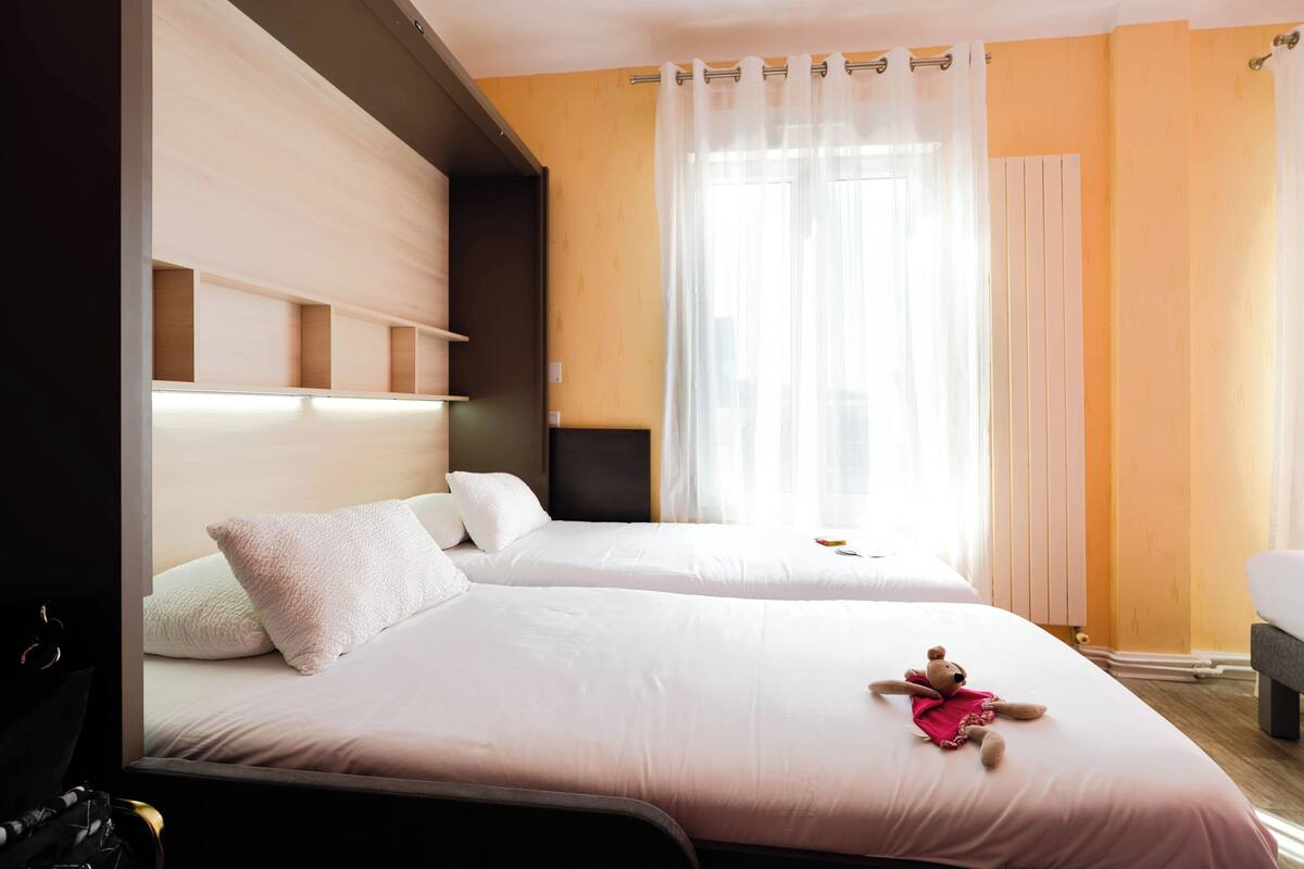 Une chambre d’hôtel avec deux lits, une tête de lit, une fenêtre avec des rideaux légers et une petite table.