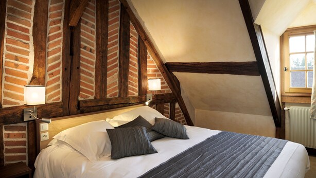Une chambre avec des poutres apparentes, un mur en briques, un lit recouvert de draps blancs et agrémenté d’oreillers gris, et une fenêtre donnant sur l’extérieur.