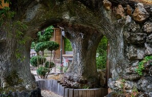 Un arc en pierre rustique mène à une construction en bois donnant sur un jardin.