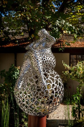 A sculpture of a woman made of reflective discs, placed outdoors with a building and greenery in the background. - La Breche (Amboise)
