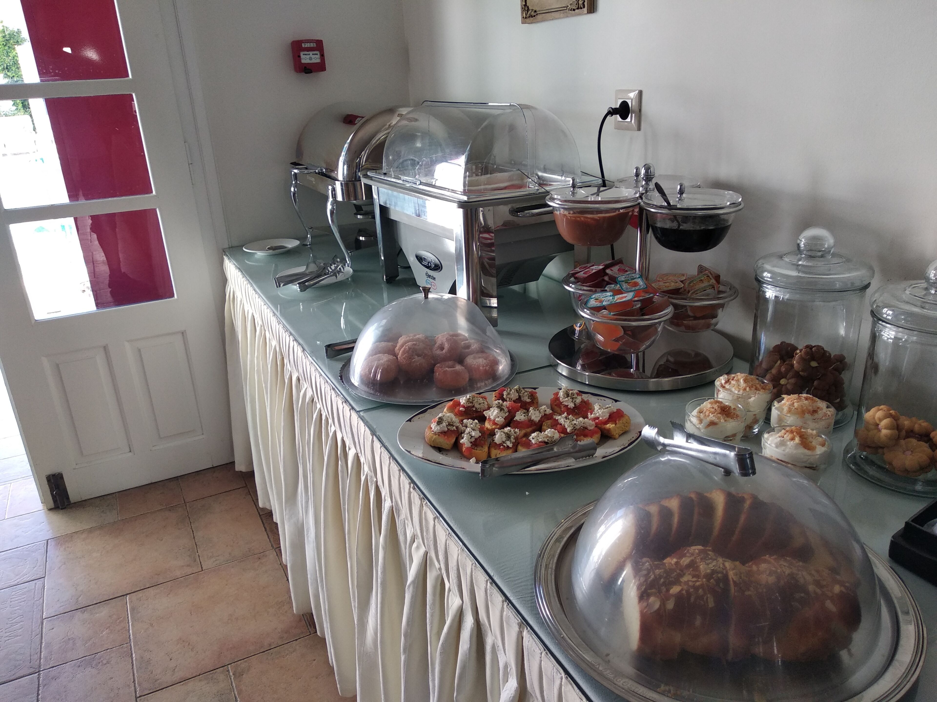 Montaje de bufé con diferentes opciones de comida, incluyendo pasteles, sándwiches y postres, cubiertos con campanas de plástico transparente.