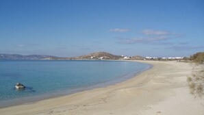 A sandy beach with clear blue water, a small rock in the water, and a distant hill with buildings. - Hotel Katerina (Naxos)