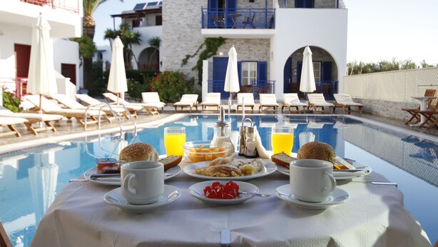A poolside breakfast table with a variety of food items and drinks, surrounded by lounge chairs and umbrellas, with a pool and buildings in the background. - Hotel Katerina (Naxos)