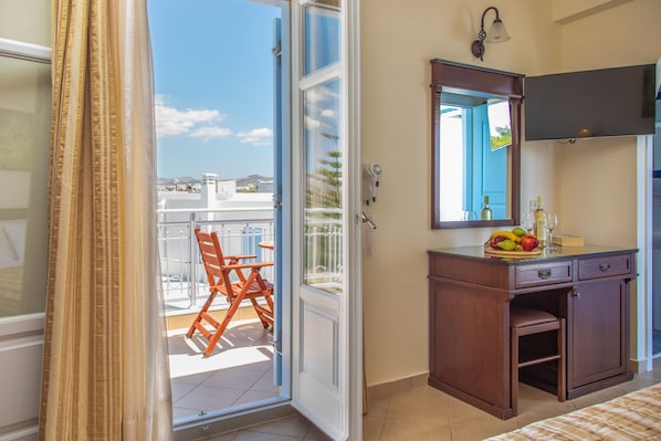 A room with a wooden desk, a mirror, and a view of a balcony with a chair. - Hotel Katerina (Naxos)