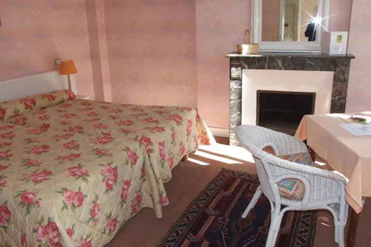 Ein Schlafzimmer mit floralem Bettbezug, einem weißen Stuhl, einem kleinen Tisch und einem Kamin.