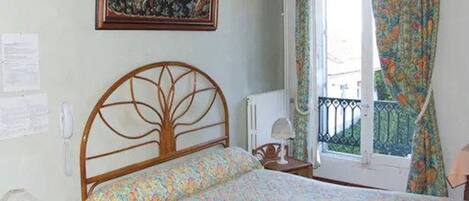 Un dormitorio con estructura de cama de madera, ropa de cama floral, un cuadro en la pared y una ventana con cortinas estampadas.