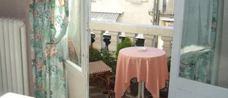 Een kamer met een bed, een raam met bloemen gordijnen, een klein balkon met een tafel en stoelen, en een geruit vloer.