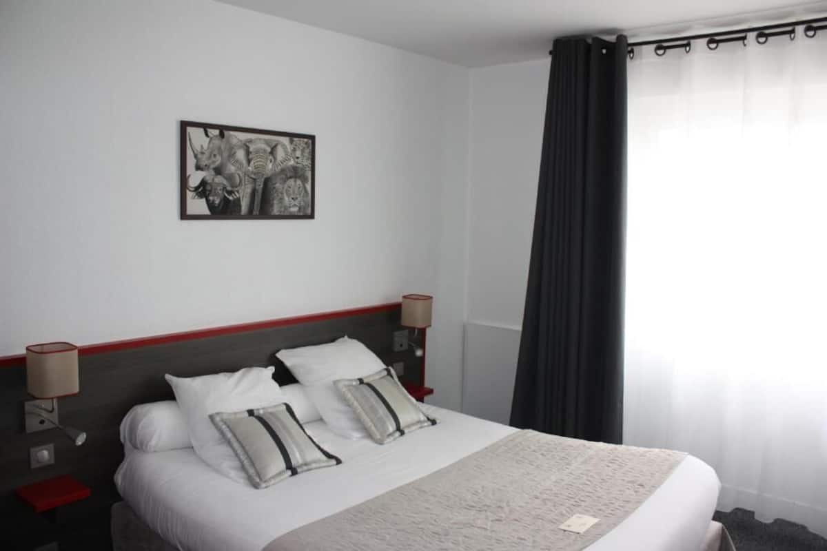 Habitación de hotel con una cama, dos mesitas de noche, una lámpara, un cuadro en la pared y una ventana con cortinas.