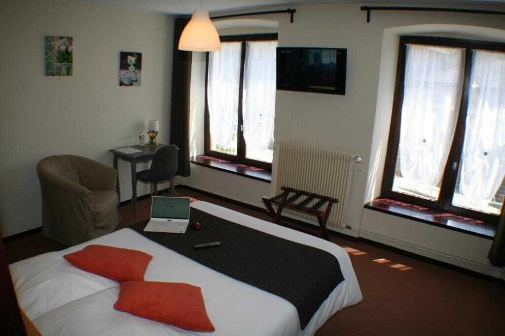 A hotel room with a bed, a desk with a laptop, a chair, and two windows with curtains.