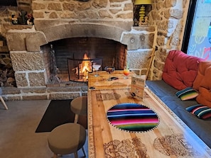 A cozy room with a stone fireplace, a wooden table with a colorful cushion, and a comfortable sofa with pillows. - Hôtel Restaurant Le Barriol (Saint-Julien-Chapteuil)
