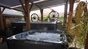 A hot tub with a wooden structure overhead, surrounded by potted plants and a stone wall in the background.