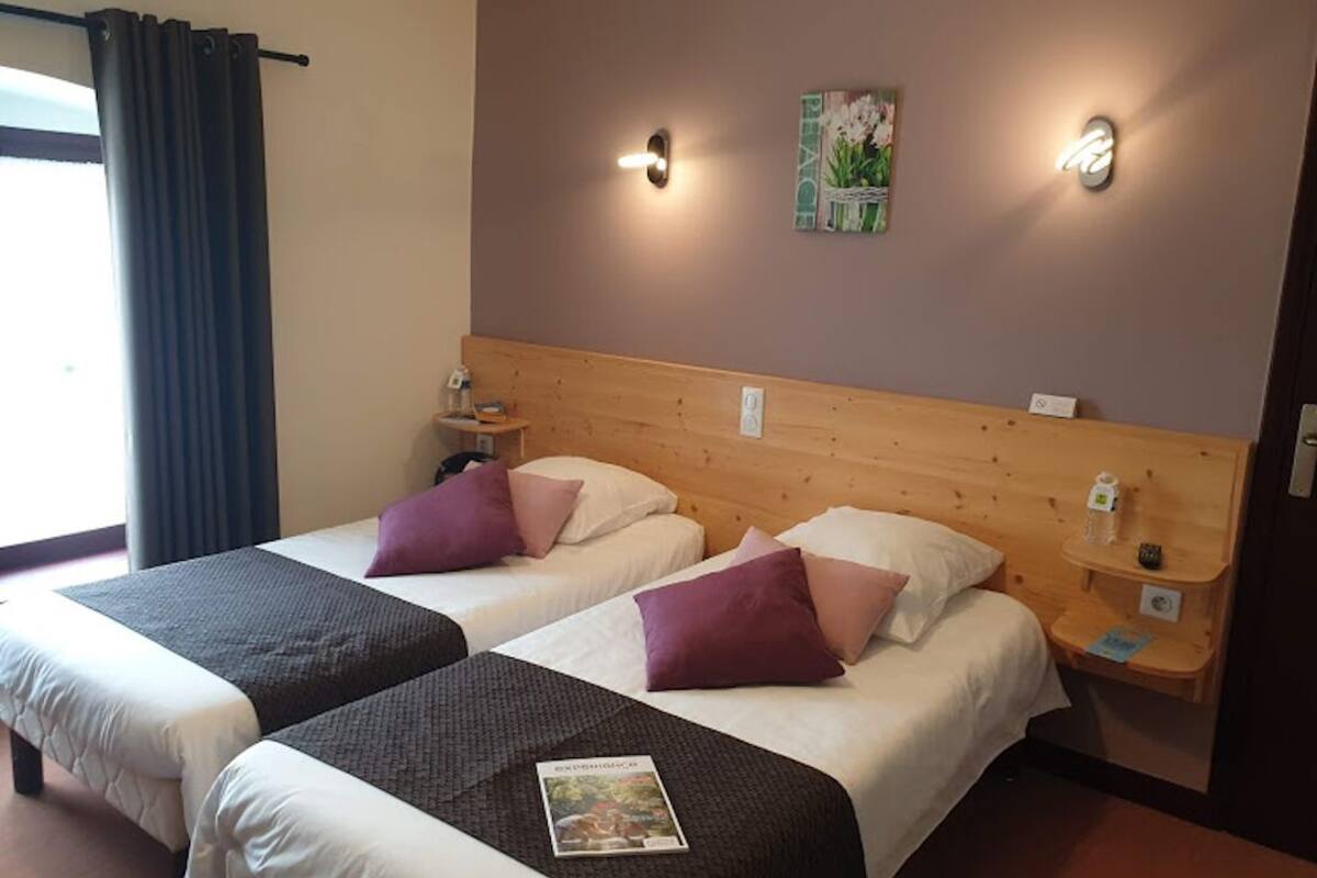 Habitación de hotel con dos camas, cada una con almohada y manta. Hay luminarias fijadas a la pared y una pequeña mesa con una botella y una taza.
