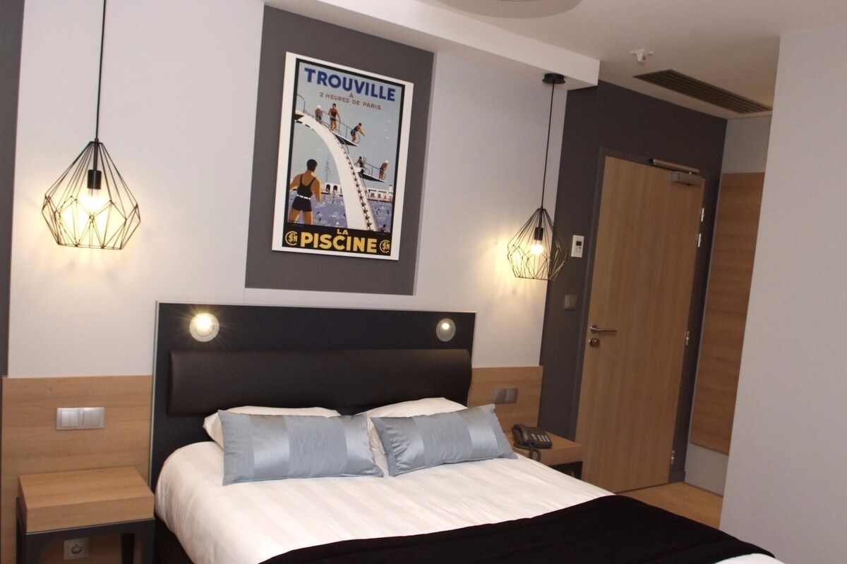 Habitación de hotel con cama, mesitas de noche, un cartel en la pared y lámparas colgantes.