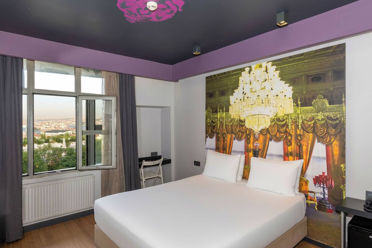 A hotel room with a large bed, a desk, and a view of the city through the window.