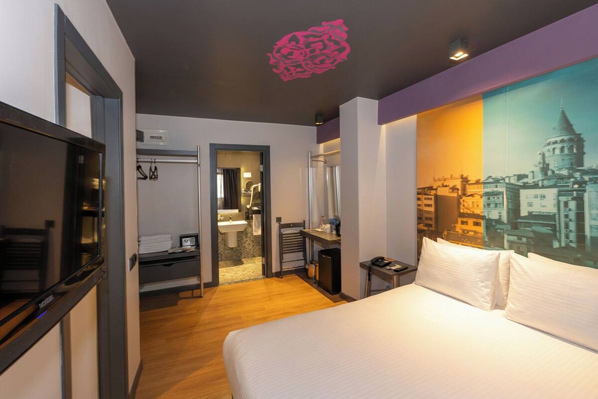 A hotel room with a bed, a desk, a mirror, a door leading to a bathroom, and a cityscape painting on the wall.
