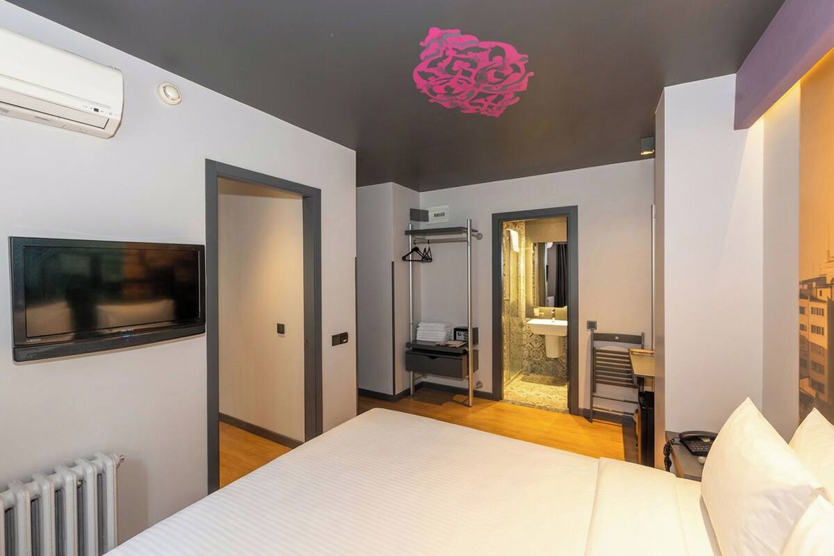 A modern hotel room with a large bed, a flat-screen TV, a wardrobe, and a bathroom with a mirror and towel rack.