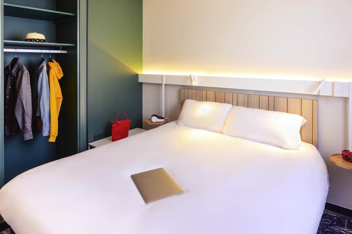 A neatly made bed with white linens, a laptop on the bed, and a red handbag on the nightstand.