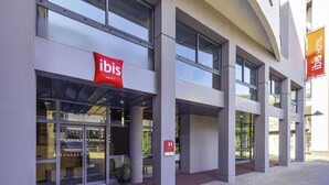 Une entrée d’hôtel moderne avec une façade en verre, une enseigne rouge « Ibis Hotels » et un symbole indiquant l’accessibilité aux personnes à mobilité réduite.