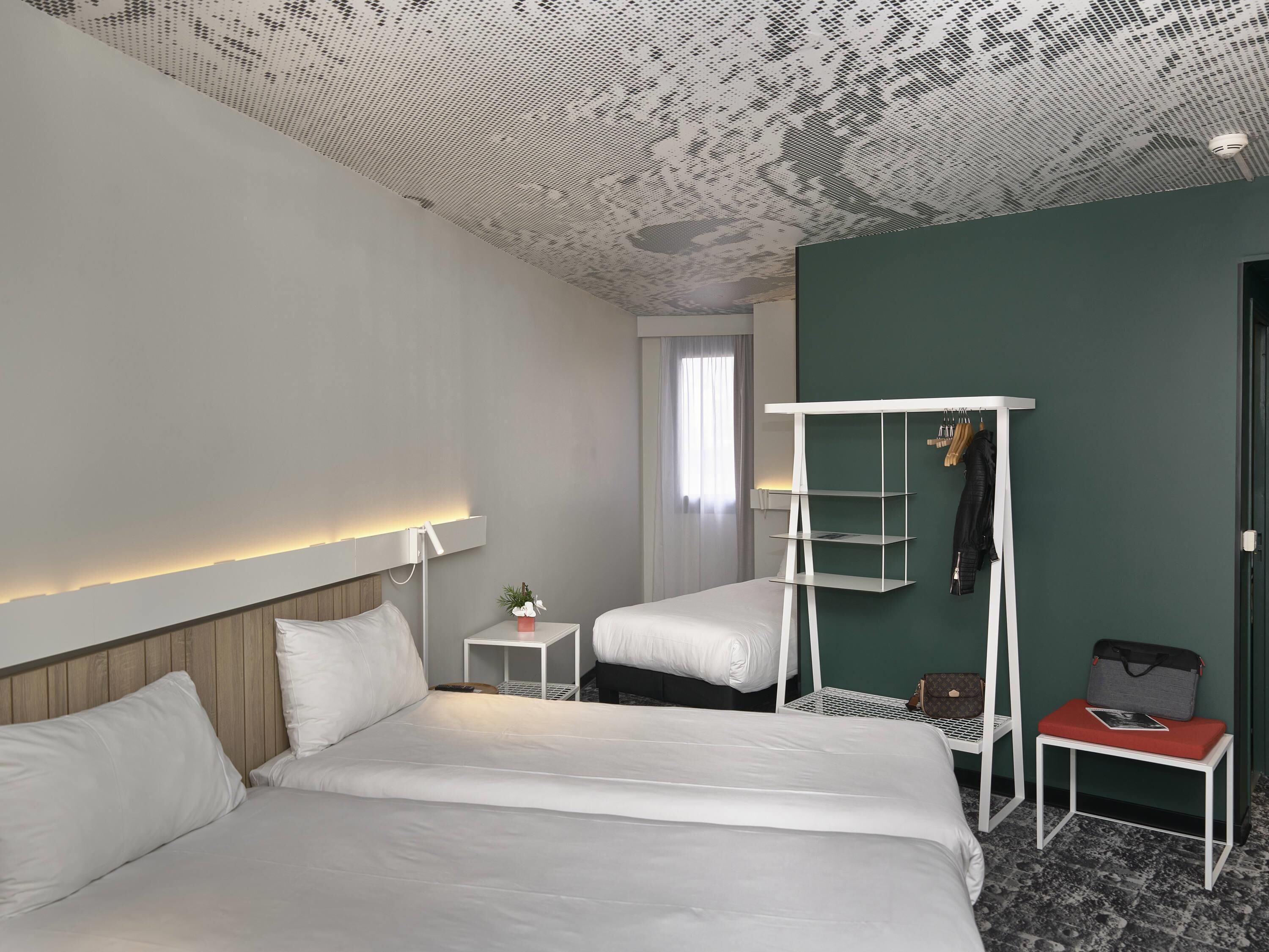 Ein Hotelzimmer mit zwei Betten, einem weißen Nachttisch mit Lampe, einem kleinen Tisch mit Pflanze und einem Kleiderständer mit Jacke und Tasche.
